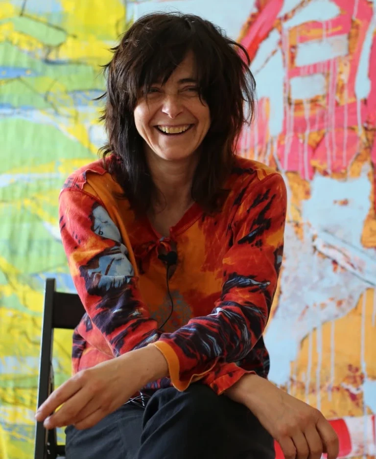 Monika Ślósarczyk seated and smiling with two colourful paintings behind her.